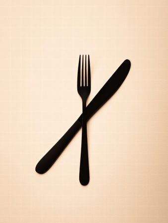 A black fork and knife are crossed on a soft beige background, highlighting minimalism in dining utensils and design simplicity.の素材