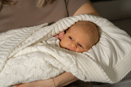 A mother gently cradles her infant child wrapped in a cozy white blanket while sitting on a couch,...
