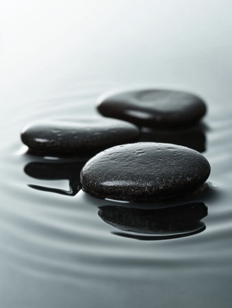 Smooth black river stones are arranged on a reflective wet surface, illuminated by soft light, highlighting their texture and creating a serene atmosphere.の素材
