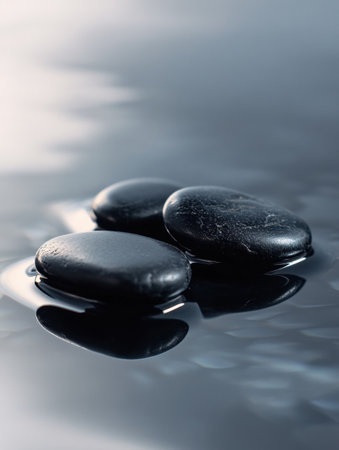 Three smooth black river stones rest on a wet reflective surface, illuminated by soft lighting that enhances their deep contrast and minimalist beauty.の素材