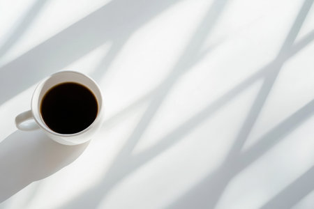 A coffee cup rests on a pristine white table, casting soft shadows that enhance the minimalist elegance while inviting a moment of calm reflection.の素材