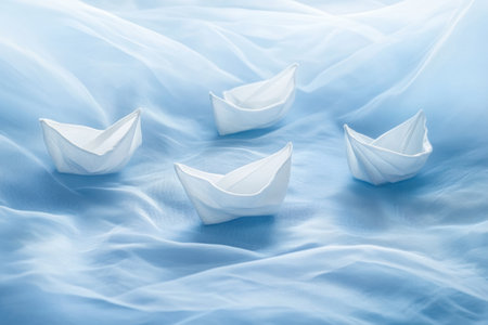Delicate paper boats drift serenely across a soft pale blue fabric, evoking a sense of calm and simplicity in a poetic arrangement of minimalist art.の素材