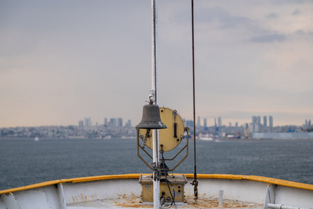 Waves gently lap against the bow of the ship as a bell hangs above, offering a view of the city skyline along the Bosphorus strait on a cloudy day.の写真素材