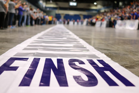 Participants cheer as runners cross the finish line marked by a large banner at a lively community event held in an indoor arena.の素材