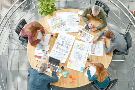 Group collaborates at round table, using devices and papers to generate ideas, while discussing plans and projects in a creative environment.の素材