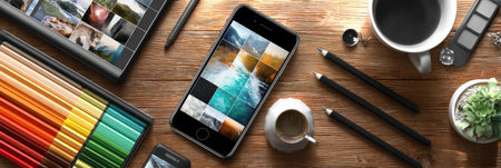 Smartphone featuring a photo editing interface is placed on a wooden desk surrounded by colorful art supplies, coffee, and a small plant, evoking creativity.の素材