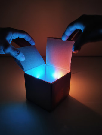 Hands joyfully open a box, revealing captivating glowing lights that illuminate the surroundings with vibrant colors and excitement during an unboxing event.の素材