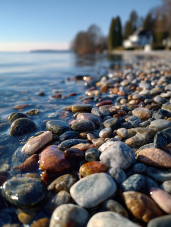 Smooth pebbles bask under the sun along a tranquil lakeside, gently touched by water and framed by distant trees on a beautiful day.の素材