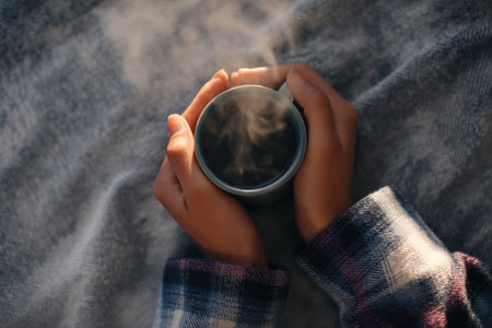 Hands hold gently a steaming cup, surrounded by a soft flannel blanket, creating a warm and inviting atmosphere on a cool morning.の素材