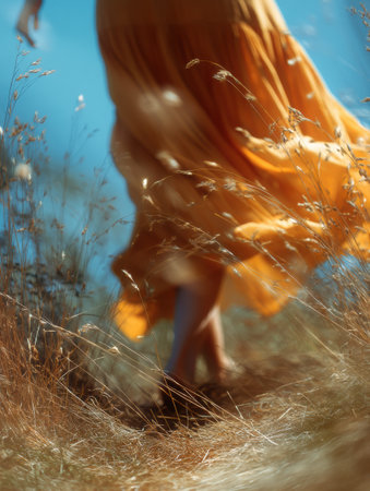 A woman in a flowing summer dress walks through golden grass, her movement creating a blur against the vibrant blue sky on a sunny day.の素材