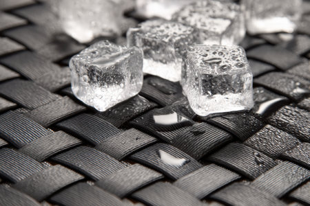 Ice cubes gradually melt on a textured woven mat, leaving behind glistening droplets in a warm and inviting setting during daylight.の素材
