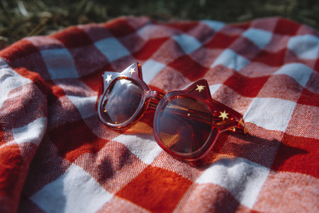 Bright star-shaped sunglasses left on a red and white checkered picnic blanket at a vibrant outdoor gathering under the warm sun in the park.の素材