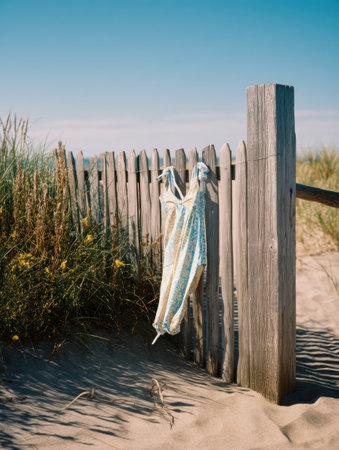A vibrant swimsuit hangs on a rustic wooden fence, swaying slightly in the warm breeze under a clear blue sky near sandy beach grass.の素材