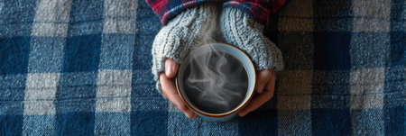Hands in knitted gloves hold a steaming cup, surrounded by a soft checkered flannel blanket, evoking warmth and comfort on a chilly day.の素材