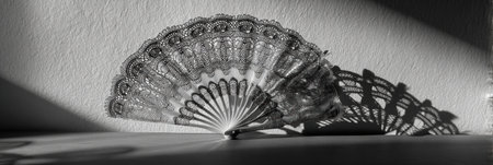 A beautifully designed paper fan rests on a surface, creating intricate shadows against a textured wall as soft light enhances its details and patterns.の素材