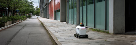A smart delivery robot moves along a deserted sidewalk lined with greenery and contemporary buildings, operating independently in broad daylight.の素材