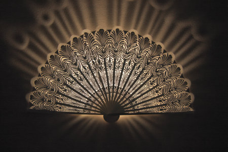 A beautifully crafted paper fan displays intricate patterns while casting complex shadows on a surface illuminated by soft, warm light.の素材