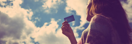 A woman stands outdoors, holding a discount card up to the sky, surrounded by soft shadows and clouds on a bright day, enjoying her moment in the sun.の素材
