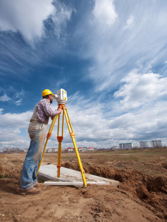 Professional surveyor stands at a construction site, adjusting a measuring tripod while surveying the land under a partly cloudy sky.の素材