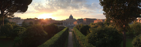 Spectacular sunrise illuminates the Vatican, with vibrant colors cascading above historic buildings, viewed through lush garden hedges in peaceful tranquility.の素材