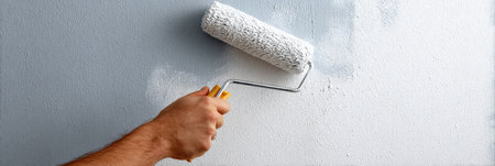 A worker is painting a wall with a roller tool, leaving a clear margin on the left side to allow for precise detailing and finishing in a bright interior area.の素材