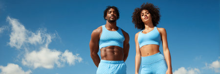 Active couple in bright blue activewear stands confidently against a sunny sky, showing their commitment to fitness and partnership in healthy living.の素材