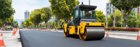 Road construction project is ongoing in a city with a roller machine compacting asphalt. Trees and buildings provide a green backdrop and urban context.の素材