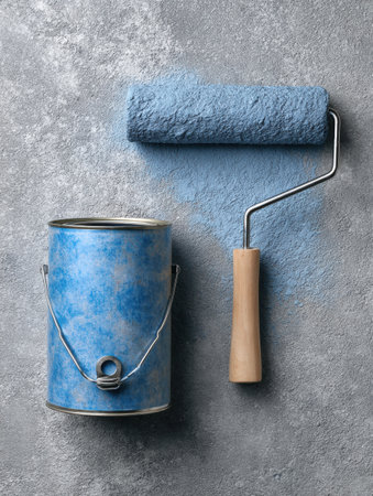 A blue paint can and roller are placed on a concrete surface, indicating preparation for a painting project or renovation task in a work area.の素材