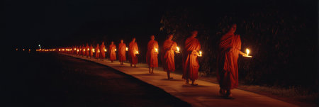 Buddhist monks in orange robes carry candles along a quiet path at night, creating a tranquil atmosphere as they practice meditation and mindfulness.の素材