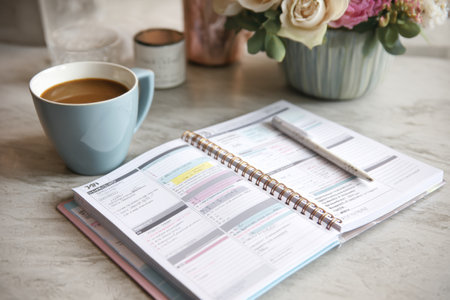 Caffeine-free daily routine planner is open on a marble table with a coffee cup, elegant flowers, and a pen nearby, creating a serene work setting.の素材