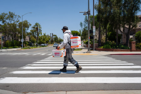 A delivery worker walks through a crosswalk with a grocery crate in a suburban neighborhood under clear blue skies, focused on completing the task.の素材
