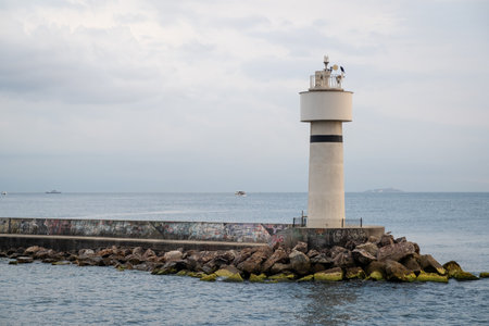 A lighthouse located on a rocky pier by the Bosphorus serves as a guiding point for passing boats, surrounded by calm waters and a dreary sky.の写真素材