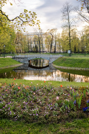 A calming view features a stone bridge over still water, vibrant flowers in front, and lush greenery framing the peaceful spring scene.の写真素材