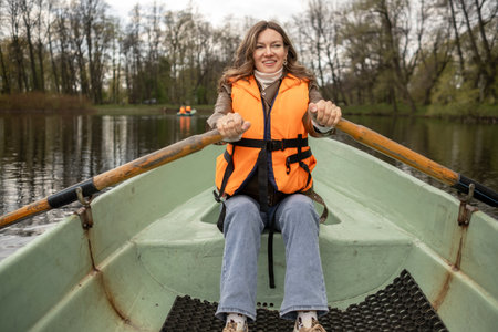 A smiling individual in a bright orange life jacket rows a small boat on a serene lake, with trees reflecting on the waters surface.の写真素材