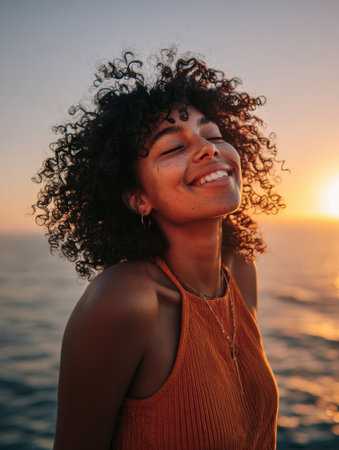 A woman with curly hair enjoys the warmth of a sunset, showing the smile that contributes to overall well-being and positive mental health benefits.の素材