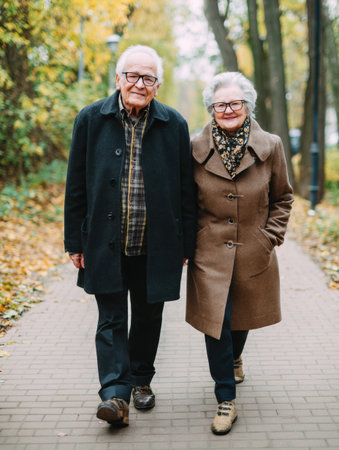 An elderly couple wearing glasses strolls hand in hand through a park, surrounded by vibrant autumn foliage and paths lined with fallen leaves.の素材