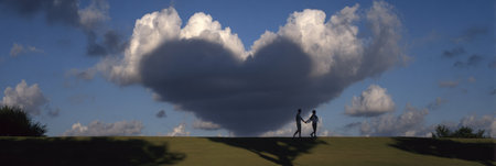 Two people stroll hand in hand through a park, casting a heart-shaped shadow with clouds above them, capturing a romantic moment at dusk.の素材