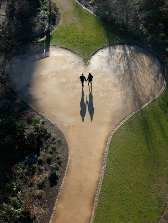 A couple strolls through a park, holding hands while their shadows form a heart shape on the pathway, with greenery surrounding them.の素材