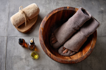 A soothing foot soak setup featuring a wooden basin, gently used towels, and an arrangement of natural oils for a calming home spa experience.の素材
