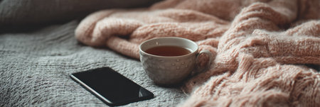 A soft blanket lies next to a cup of warm tea, while a switched-off phone rests nearby in a tranquil and cozy environment.の素材