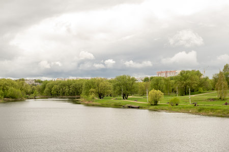 A serene river flows through a vibrant park filled with lush trees, surrounded by gently rolling land under an overcast sky in the spring season.の写真素材