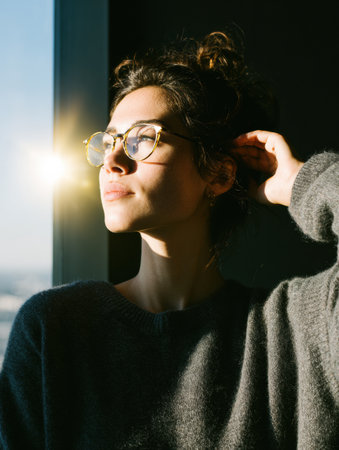 A woman stands near a window, holding eyeglasses and gazing thoughtfully as soft daylight casts a warm glow on her face, creating a peaceful atmosphere.の素材