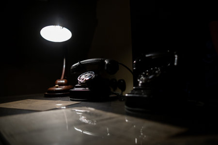Dark ambiance highlights antique telephones on a desk accompanied by a warm lamp, evoking a sense of...