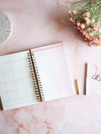 A health planner is opened on a light pink desk, providing organized space for notes, next to a small vase of flowers and writing tools.の素材