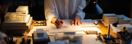 Person focused on sketching an architectural model at a desk surrounded by materials and warm lighting, creating an inviting atmosphere for design work.の素材