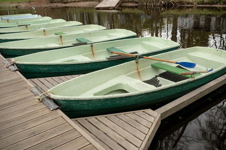 Several green rowboats are lined up against a wooden dock, reflecting the tranquil river and surrounding trees under cloudy skies.の写真素材