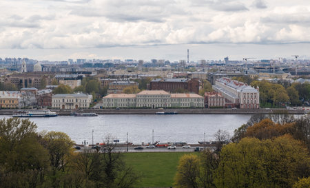 View of notable architecture along the river in St Petersburg on a cloudy day, highlighting the blend of nature and urban life with green trees.の写真素材