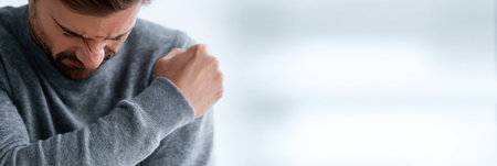 A man sneezes into his elbow, promoting hygiene and public health in a clean, focused setting.の素材