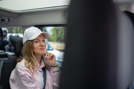 A woman with long hair sits in a car, wearing a cap and a pink shirt, reflecting thoughtfully as she enjoys the journey in the daylight.の写真素材