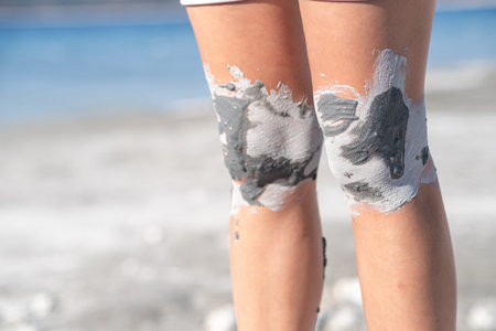 A person stands on the beach, covering their knees with natural clay while the sun shines and waves...